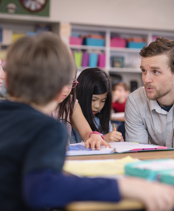 Ein junger Lehrer unterhält sich  mit zwei Schülern. Sie sitzen zu dritt an einem Tisch in einer Bibliothek. 