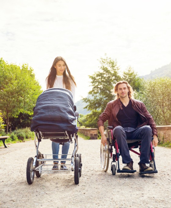 Eine Frau schiebt einen Kinderwagen, während ein Mann daneben in einem Rollstuhl sitzt. Sie spazieren gemeinsam einen Weg entlang. 