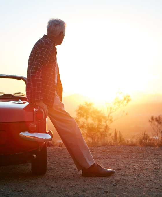 Ein erwachsener Mann lehnt an seinem Oldtimer und schaut von einem Hügel aus in den Sonnenuntergang. 