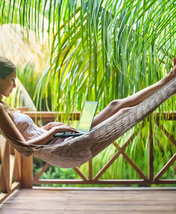 Eine Frau liegt unter Palmen in einer Hängematte mit ihrem Laptop.