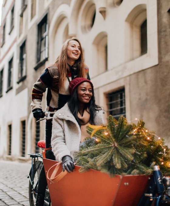 Zwei Freundinnen sind zusammen mit einem Lastenrad durch die Straßen unterwegs. Eine Person fährt das Rad, die andere Person sitzt drin. Sie transportieren einen kleinen Nadelbaum.