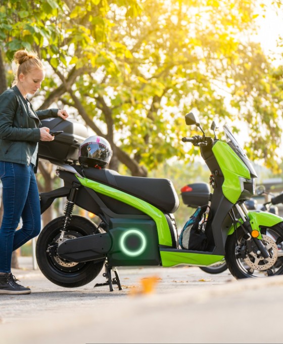 Eine blonde Frau steht neben ihrem elektrischen Motorrad und hält ihren Helm in der Hand. 