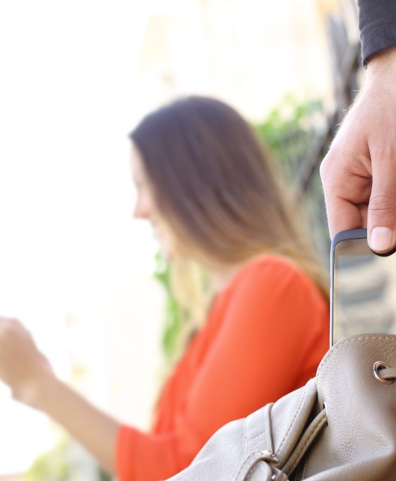 Eine Frau sitzt auf einer Parkbank und liest etwas, während ihr aus der Handtasche das Smartphone geklaut wird. 