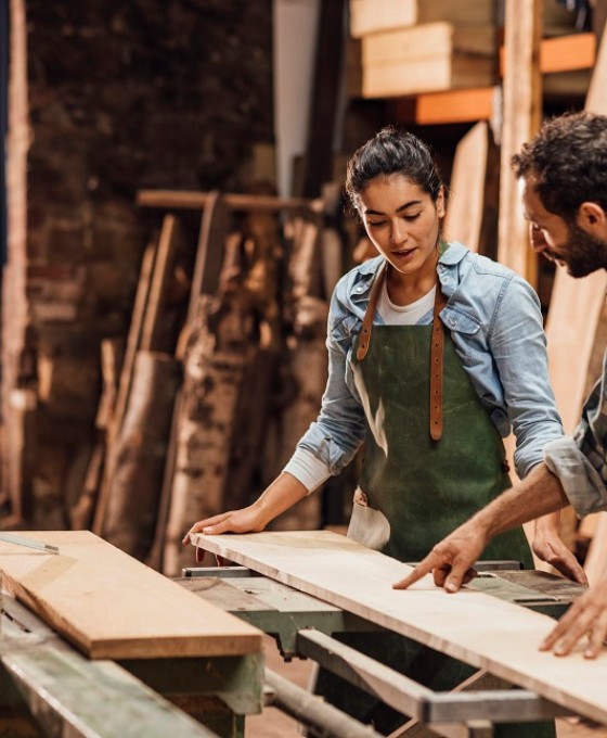 Eine junge Frau und ein junger Mann schneiden zusammen in einer Werkstatt Holz zu.