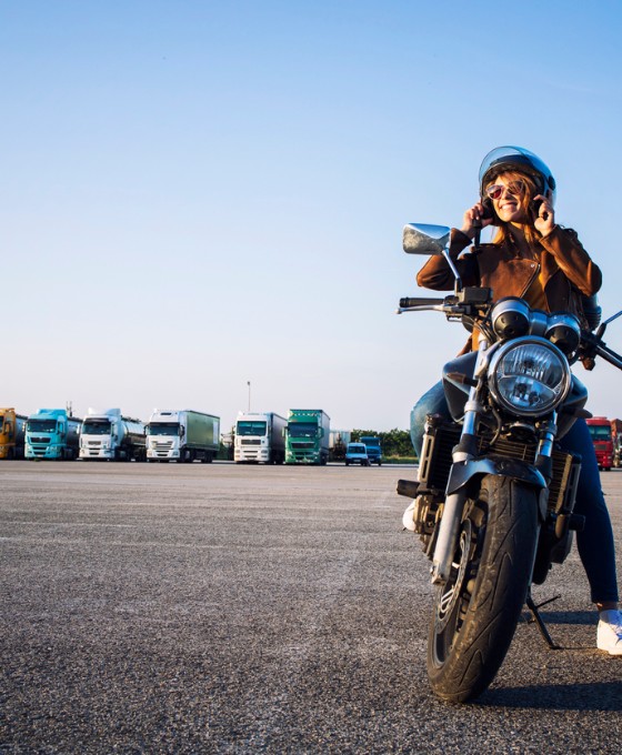 Frau sitzt mit Helm auf einem Motorrad auf einem großen Parkplatz, im Hintergrund stehen Lkw.