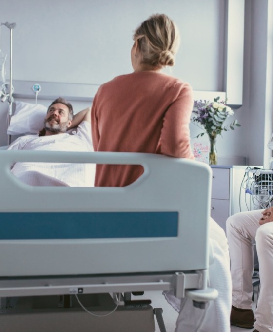 Ein männlicher Patient liegt in einem Krankenhausbett. Rechts von ihm steht eine weibliche Ärztin, eine Frau sitzt am Fußende des Bettes und ein Mann daneben auf einem Stuhl. 