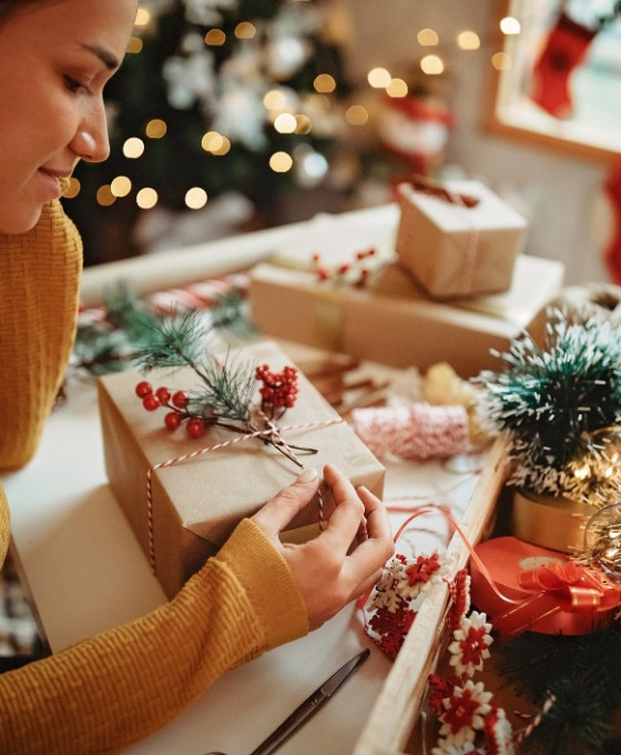 Ein Mädchen sitzt nachdenklich am Schreibtisch und begutachtet ein eingepacktes Weihnachtsgeschenk. Um sie herum ist Weihnachtsdekoration und andere eingepackte Weihnachtspäckchen. 