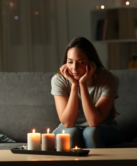Eine junge Frau sitzt in ihrem Wohnzimmer ohne Strom und schaut ernüchternd auf drei leuchtende Kerzen auf dem Wohnzimmertisch. 