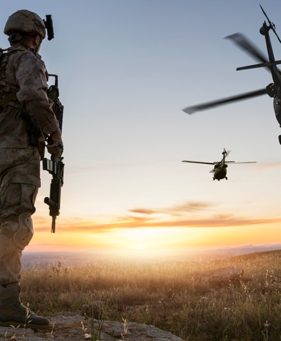 Ein Soldat steht bewaffnet auf einem Hügel schaut in Richtung wegfliegender Militärhubschrauber. 
