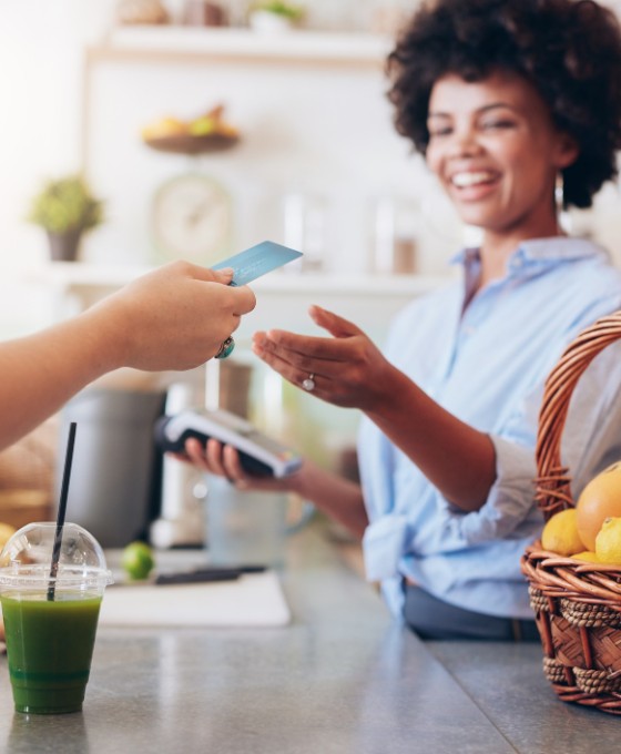 Eine Hand hält einer Frau eine Bankkarte hin. Auf dem Tresen steht ein Smoothie und die Frau hinter dem Tresen kassiert ab. 