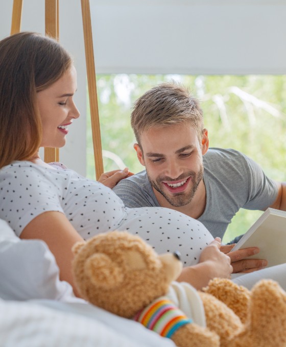 Eine schwangere Frau liegt im Bett. Ein Mann liegt neben ihr und liest ein Bett vor, während er auf den Babybauch schaut. 
