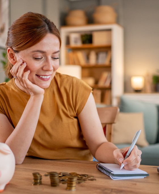 Eine junge Frau sitzt am Schreibtisch und schreibt etwas auf. Vor ihr steht ein Sparschwein.