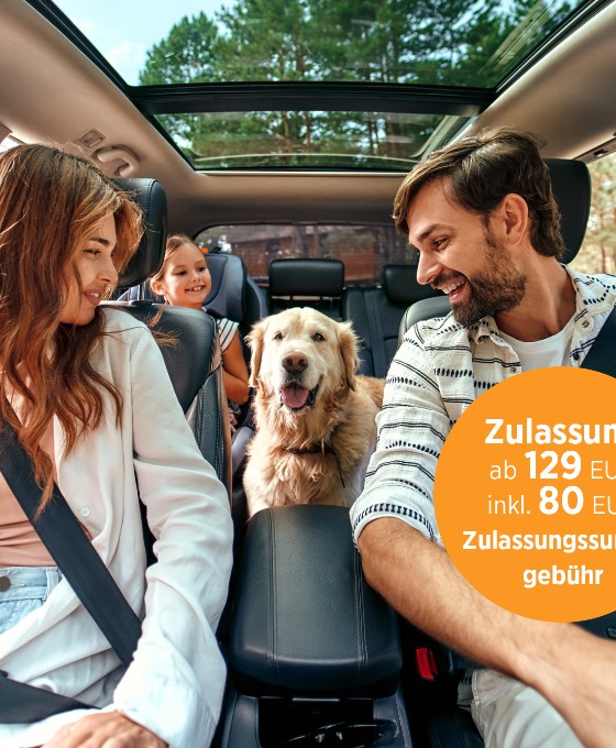 Eine junge Familie, fotografiert aus der Perspektive des Autocockpits, sitzt angeschnallt Auto und haben ihren goldbeigen Hund auf der Rückbank mit dabei.