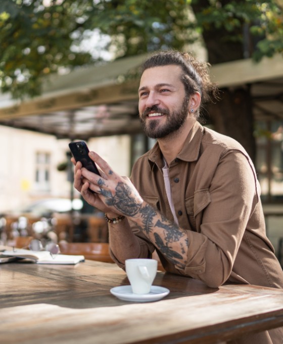 Ein Mann mit Tattoos am Arm sitzt im Außenbereich eines Cafés, hat eine Tasse vor sich stehen und blickt lachend Richtung Straße.