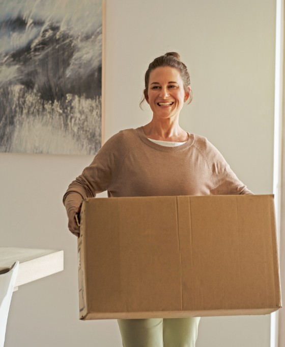 Eine Frau trägt lächelnd einen Umzugskarton durch ein Wohnzimmer.