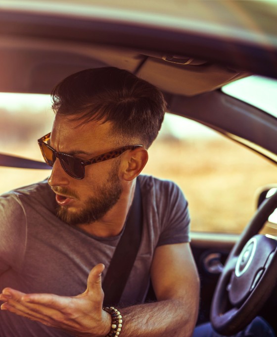 Ein Mann mit Sonnenbrille schaut verwundert und Schultern zuckend um sich im Auto.  