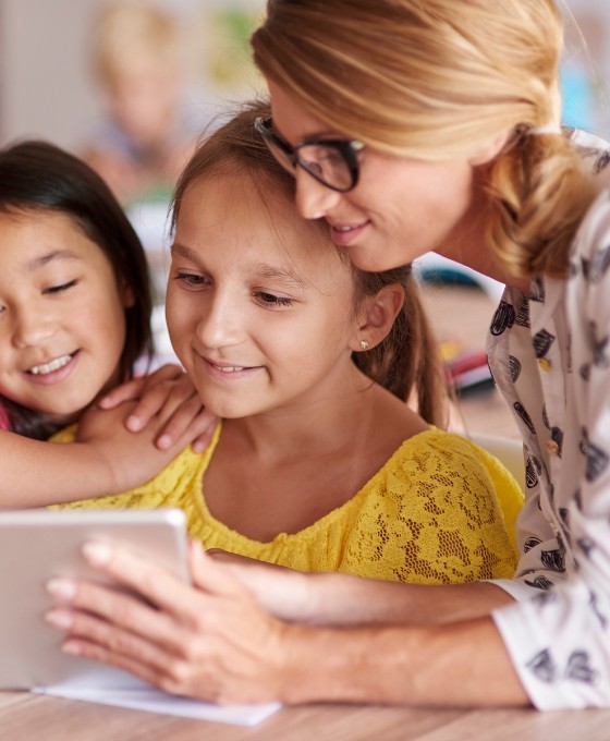 Eine Lehrerin zeigt zwei jungen Schülerinnen etwas auf dem Tablet. Sie sitzen in einem Klassenzimmer.