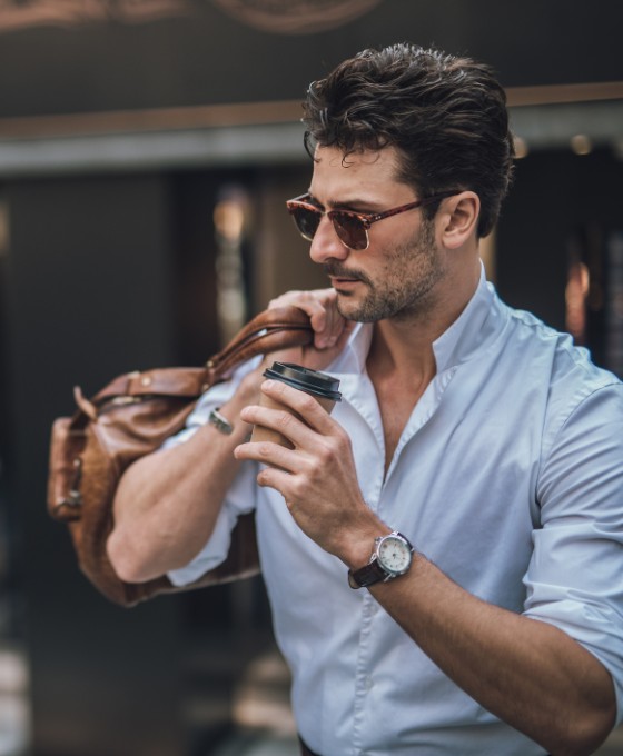 Junger Mann mit Sonnenbrille trinkt einen Kaffee während er seine Reisetasche über eine Schulter sowie eine Luxusuhr trägt. 