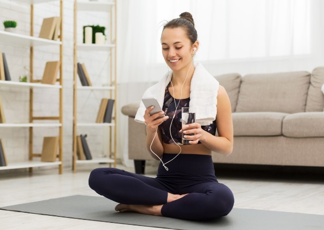 Eine Frau sitzt im Schneidersitz auf einer Sportmatte und lacht in ihr Handy.