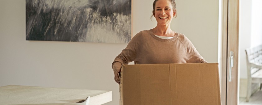 Eine Frau trägt lächelnd einen Umzugskarton durch ein Wohnzimmer.