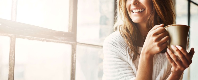 Eine lächelnde Frau sitzt am Fenster mit einer Tasse in der Hand.