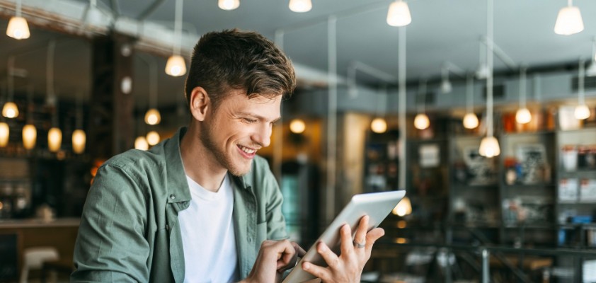 Junger Mann mit grünem Hemd liest in einem Bücher-Cafe auf einem Tablet und lächelt