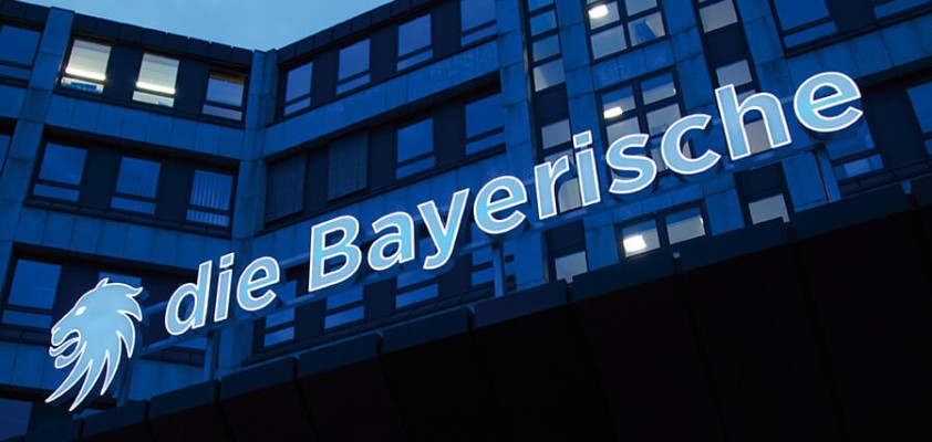 Eingang eines Bürogebäudes mit dem beleuchteten Schriftzug "die Bayerische" bei Dämmerung.