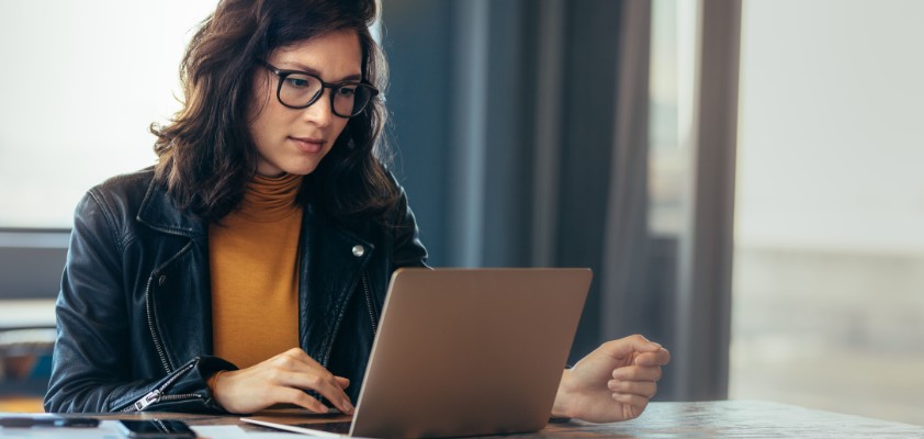 Eine Frau sitzt an einem Tisch und schaut auf ihren Laptop. Neben ihr liegen Dokumente. 