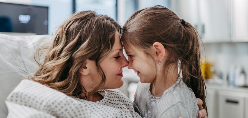 Eine Frau im Krankenbett und ihre kleine Tochter geben sich einen Nasenkuss