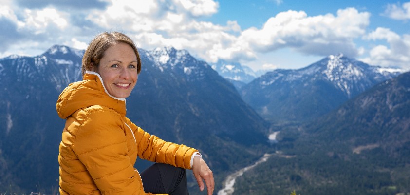 Magdalena Neuner sitzt auf einer Wiese in den Bergen und blickt in die Kamera.