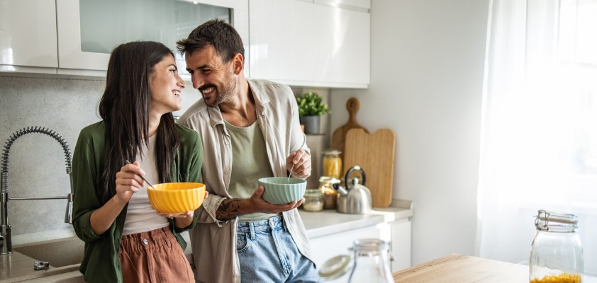 Ein junges Paar steht mit Müslischalen in der Hand steht lachend in der Küche