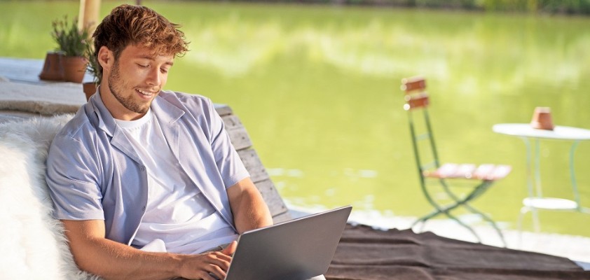 Junger Mann mit T-Shirt und Sommerhemd sitzt an einem See und schaut in ein Laptop