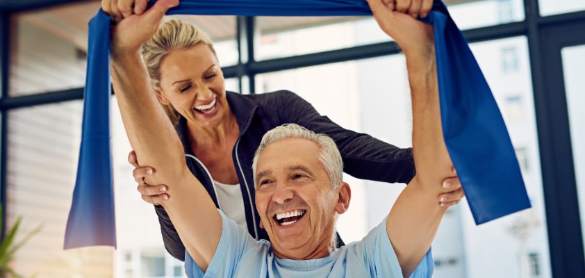 Ein glücklicher, älterer Herr führt mit einem Band eine Physioübung aus, während seine Physiotherapeutin ihn im Hintergrund stützt. 