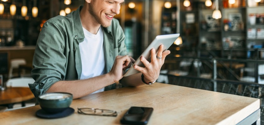 Junger Mann mit grünem Hemd liest in einem Bücher-Cafe auf einem Tablet und lächelt