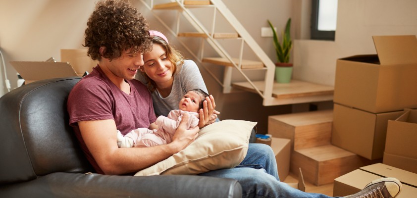 Junges Pärchen mit Baby im Arm sitzen im Wohnzimmer, voll mit Umzugskartons