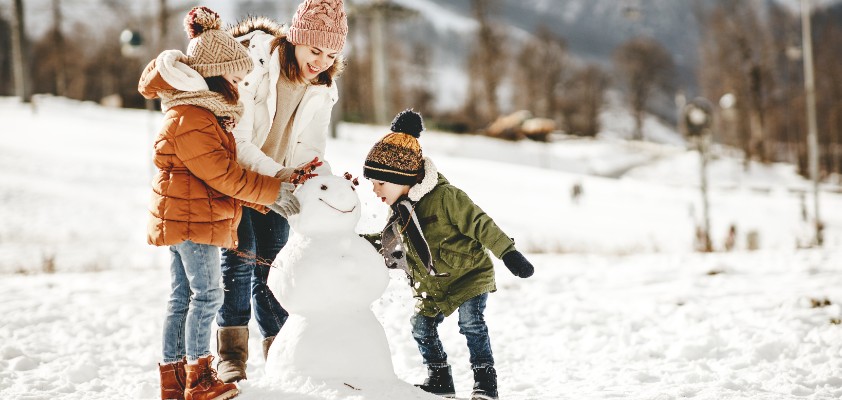 Eine Mutter baut mit ihren zwei Kindern einen Schneemann in einer Winterlandschaft. 