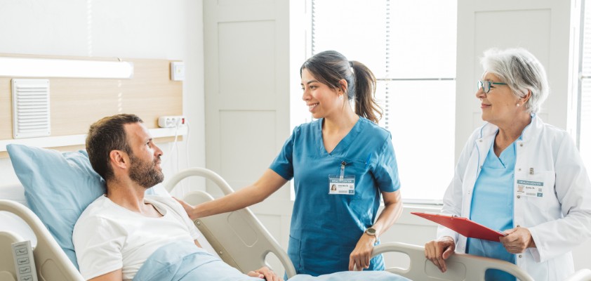 Eine junge Pflegerin und eine ältere Ärztin stehen gemeinsam an einem Patientenbett und unterhalten sich mit dem dort liegenden männlichen Patienten.