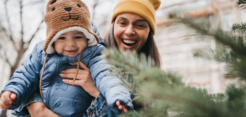 Glückliche Frau hält ihr Baby vor einem Tannenbaum.
