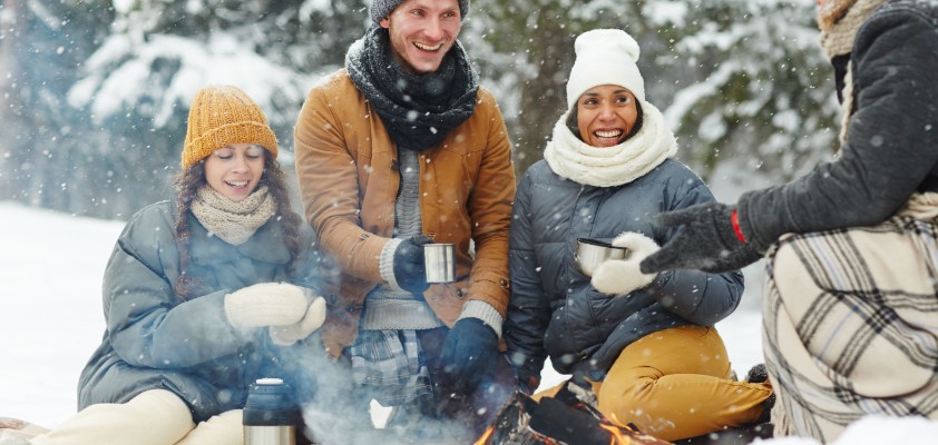 Eine Gruppe von Freunden sitzt in Winterklamotten im Schnee und grillt. 