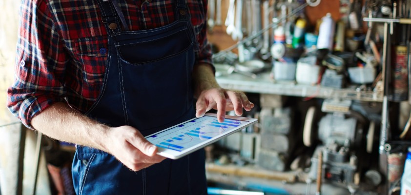 Ein Handwerker steht in einer Garage mit Handwerksequipment und schaut auf ein Tablet. 