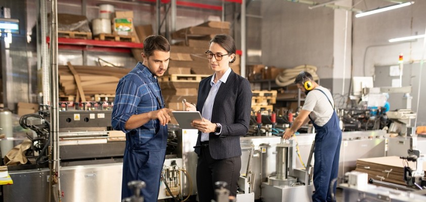 Eine Business-Frau und ein Bauarbeiter sprechen zusammen über ein Dokument, welches die Frau in der Hand hält. Die Umgebung ist eine Fabrik. 