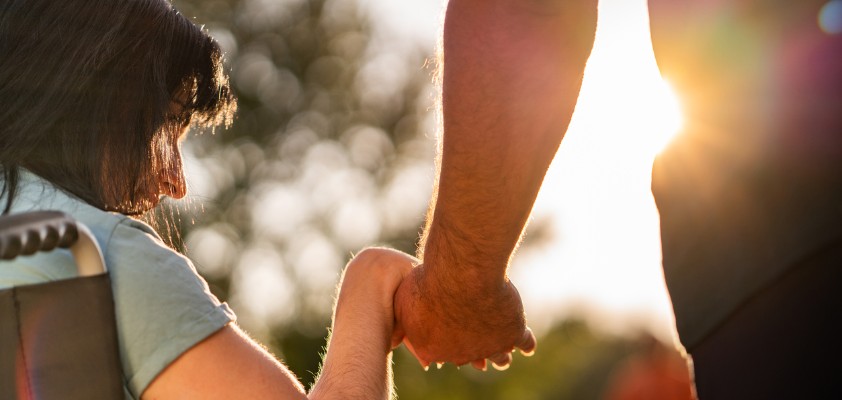 Person im Rollstuhl hält die Hand einer anderen Person im Sonnenlicht.
