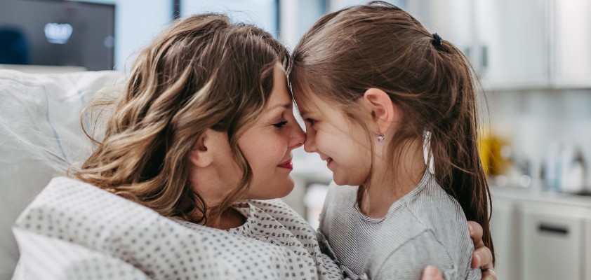 Eine Frau im Krankenbett und ihre kleine Tochter geben sich einen Nasenkuss