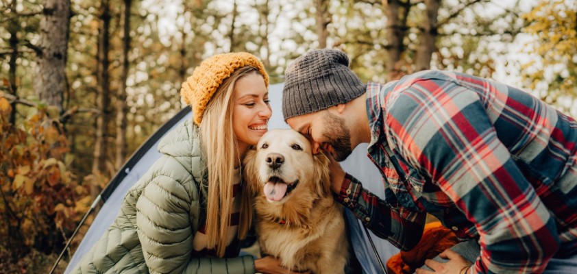 Frau mit gelber Mütze und Mann mit kariertem Hemd beim Camping mit Golden Retriever