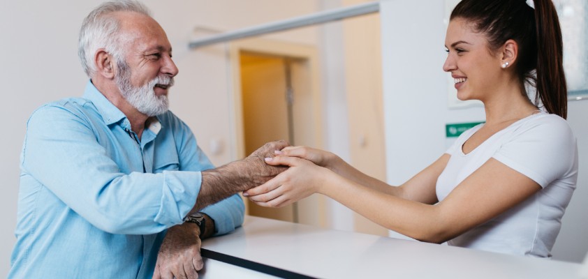 Eine junge Arzthelferin steht hinter der Empfangsrezeption und hält die Hand eines älteren Patienten, der vor ihr am Tresen steht. 