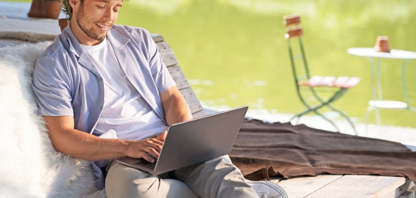 Junger Mann mit T-Shirt und Sommerhemd sitzt an einem See und schaut in ein Laptop