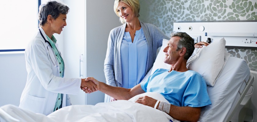 Ein Patient im Krankenhausbett schüttelt eine Ärztin die Hand, während seine Frau daneben steht.