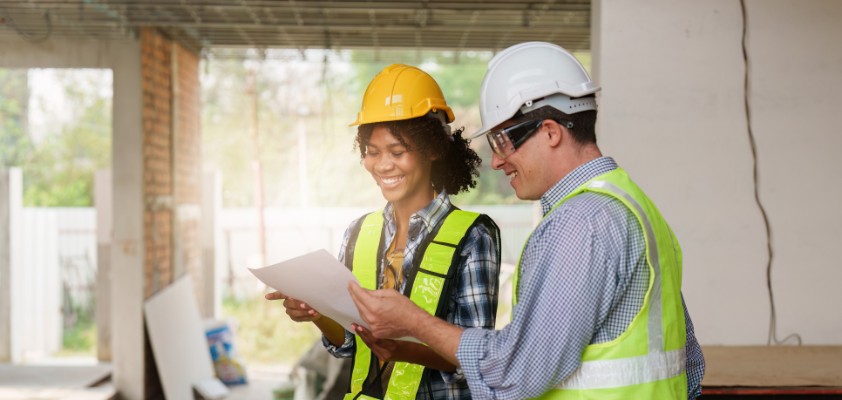 Ein Bauherr und eine Baufrau lesen sich Baupläne durch, während sie in Schutzkleidung auf einer häuslichen Baustelle stehen.