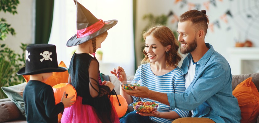 Zwei auf dem Sofa sitzende Eltern geben ihren verkleideten Kindern Süßigkeiten in ihren Halloween-Eimer. 