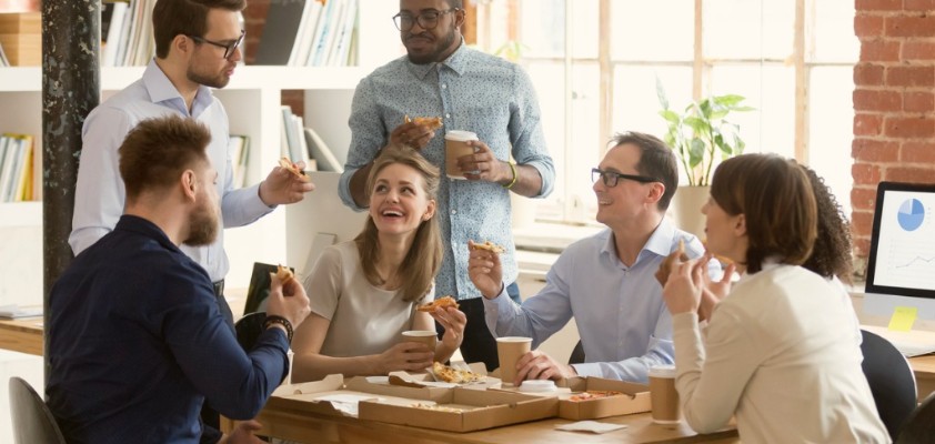Eine Gruppe von Kollegen sitzt lachend zusammen im Büro an einem Tisch und isst Pizza. 